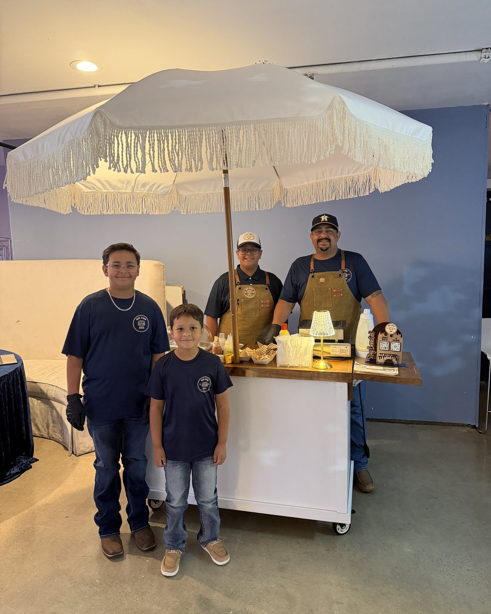 The Mini Stack Bros family in aprons at their pancake cart under a fringed white umbrella